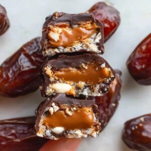 Chocolate caramel stuffed dates on a marble board, plump Medjool dates split open showing creamy nut butter and caramel filling, dipped in glossy dark chocolate, sprinkled with flaky sea salt and chopped pistachios