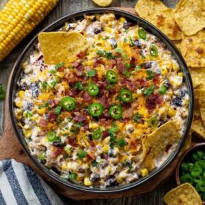 Corn dip with bacon in a cast iron skillet, golden bubbly baked dip with melted cheese on top, sweet corn kernels visible, crispy bacon crumbles and diced jalapeño scattered on surface, sliced green onions, tortilla chips and crackers arranged around the skillet