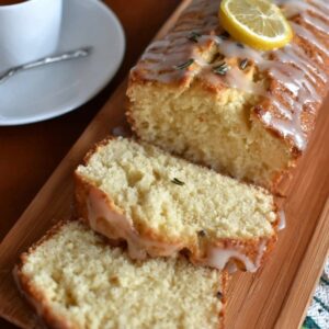 Lemon rosemary pound cake on a wooden serving board with a yellow honey-lemon glaze drizzled over the top, fresh rosemary sprigs garnishing, lemon zest curls, slice cut showing tight buttery crumb